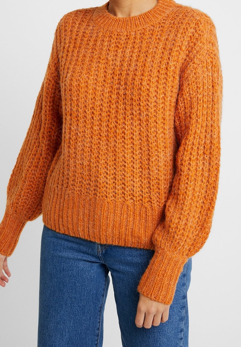 Personne portant un pull orange épais en tricot texturé à manches longues et un jean bleu sur un fond clair uni.