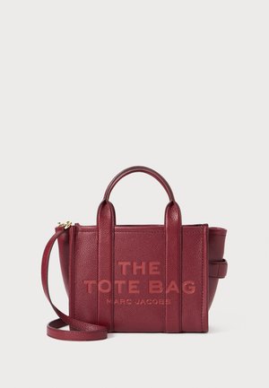 Bordeaux leren tote bag met gestructureerd oppervlak, twee korte handvatten en een verwijderbare schouderriem. Geprinte tekst aan de voorkant: "DE TOTE BAG MARC JACOBS."