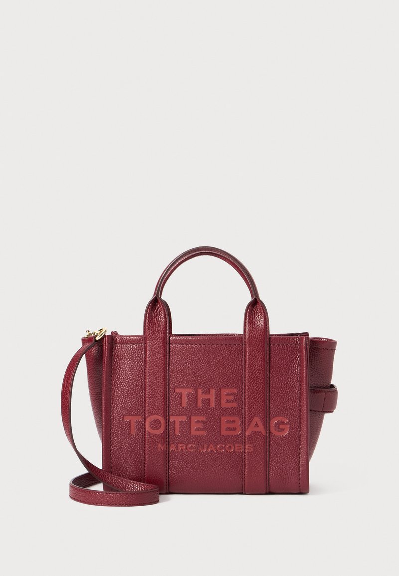 Maroon leather tote bag with textured surface, two short handles, and a removable shoulder strap. Printed text on front: "THE TOTE BAG MARC JACOBS."