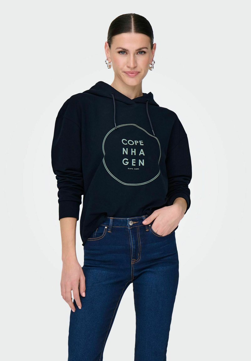 Felpa con cappuccio blu navy con una grafica circolare che riporta la scritta "COPE NHGEN," abbinata a jeans blu scuro. Presenta una vestibilità comoda e lacci.