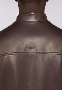 Bruine lederen jas met een gladde textuur, voorzien van een staande kraag en het geperste "TRUSSARDI" logo op de rug. Minimale stikdetails.