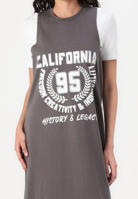 Robe grise sans manches avec imprimé graphique blanc indiquant "CALIFORNIA 95" et des motifs floraux, portée sur une chemise blanche à manches courtes. Matière en coton.