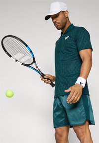 Grøn mønstret tennis polo med krave, parret med grønne satinshorts. Sort tennisraket i hånden, gul tennisbold i luften.