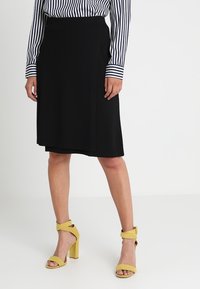 Femme portant une jupe noire à mi-longueur, une chemise à rayures noires et blanches à manches longues, et des sandales à talons hauts jaunes avec des brides autour de la cheville.