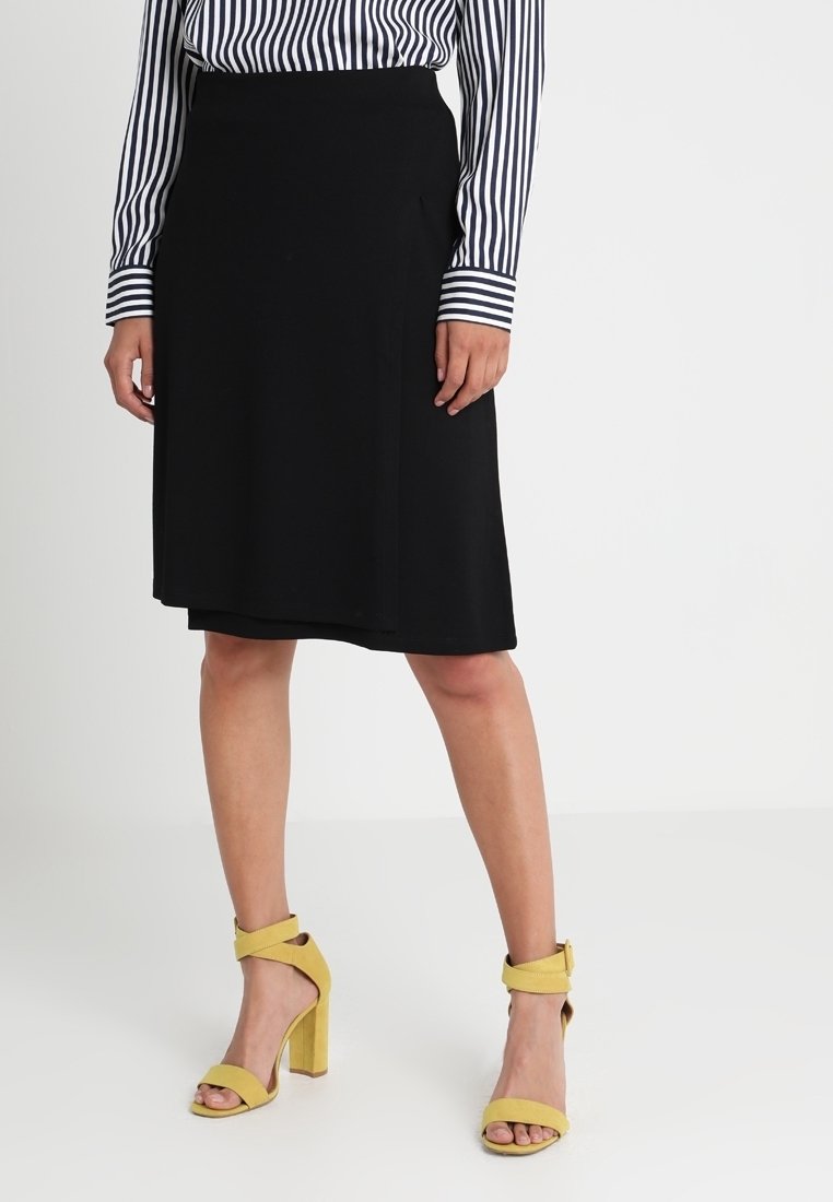 Femme portant une jupe noire à mi-longueur, une chemise à rayures noires et blanches à manches longues, et des sandales à talons hauts jaunes avec des brides autour de la cheville.