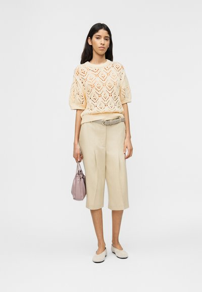 Femme portant un pull à manches courtes en maille beige, un culet beige taille haute avec ceinture, tenant un sac à main mauve et des chaussures beige à enfiler.
