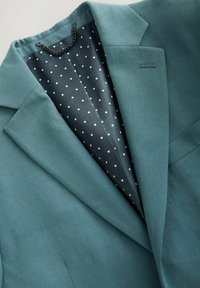 Türkes Blazer mit einer glatten Textur; das Innenfutter besteht aus einem dunklen Stoff, der mit weißen Punkten gefüttert ist, und hat ein dezentes schwarzes Kettendetail am Kragen.