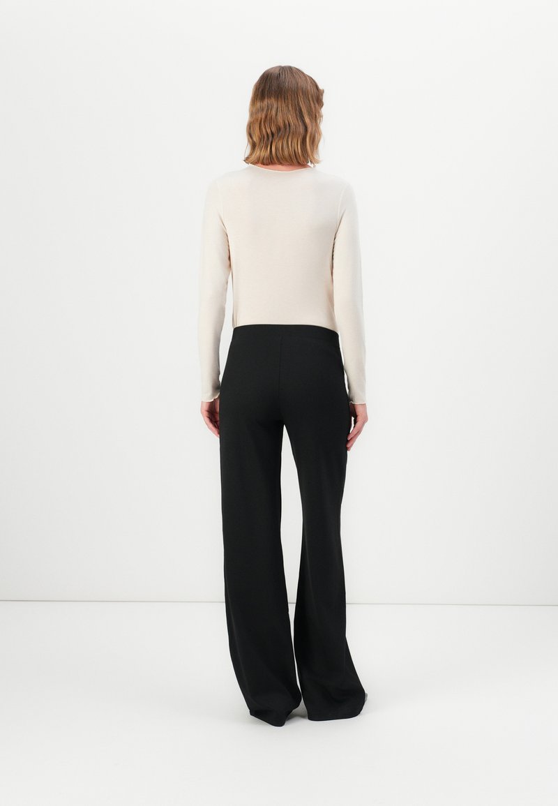 Persona con capelli ondulati di lunghezza spalla, che indossa una maglietta a maniche lunghe di colore crema e pantaloni neri larghi, in piedi di fronte a un muro bianco semplice.