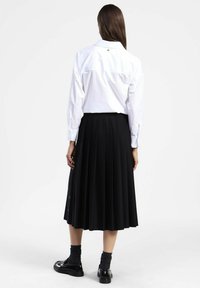 Camicia bianca con bottoni, a maniche lunghe, abbinata a una gonna nera plissettata. Tessuto liscio; la camicia presenta un colletto e una marcatura sottile.