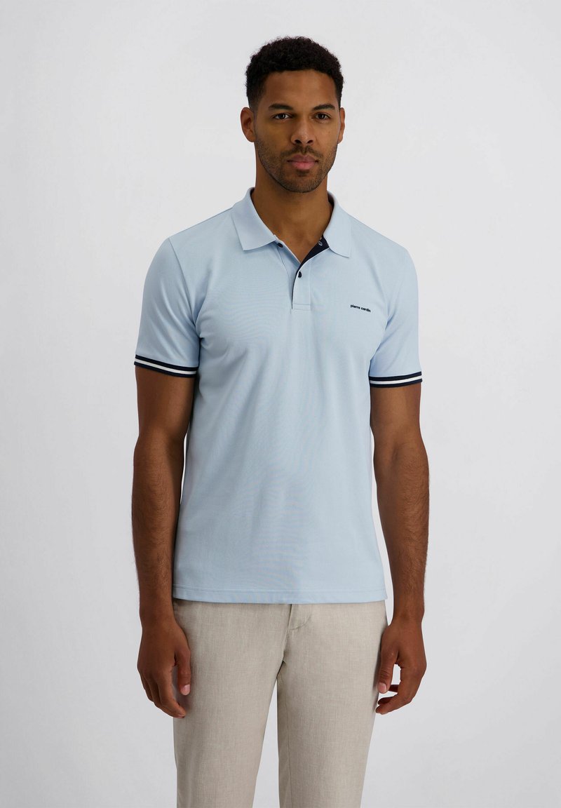 Mann trägt hellblaues Poloshirt mit schwarzen und weißen Ärmelkanten und beige Hose, steht vor einfachem Hintergrund.
