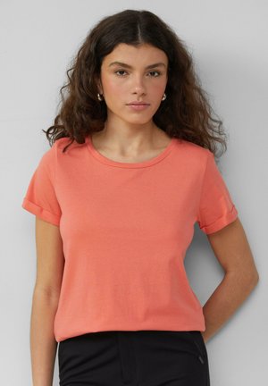 Coral short-sleeve T-shirt in soft cotton knit, featuring a crew neck and rolled sleeves, paired with dark trousers. Smooth texture and relaxed fit.