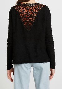 Black long-sleeve sweater with a triangular floral lace panel on the upper back, worn with light blue jeans.