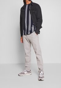 Zwarte denimjas over een marineblauw en witte gestreepte shirt, gecombineerd met lichtgrijze sweatpants en grijze sneakers met witte accenten.
