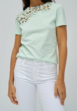 Femme portant un haut vert clair à manches courtes avec un détail en dentelle florale sur le haut de la poitrine et un pantalon blanc taille haute.
