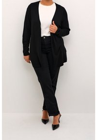 Sort langærmet cardigan med åben front, båret over en hvid top og sorte bukser, parret med sorte spidse hæle, med en afslappet pasform.