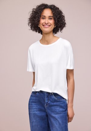 Femme souriante aux cheveux bouclés portant un t-shirt blanc uni et un jean bleu, debout devant un fond neutre.