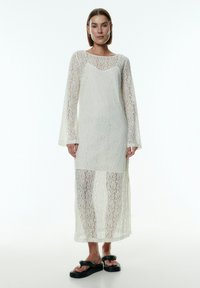 Weißes Spitzenkleid mit langen Ärmeln, transparenter Textur und hohem Ausschnitt. Verfügt über einen mittellangen Saum und ein geschichtetes Design. Schwarze Sandalen runden den Look ab.