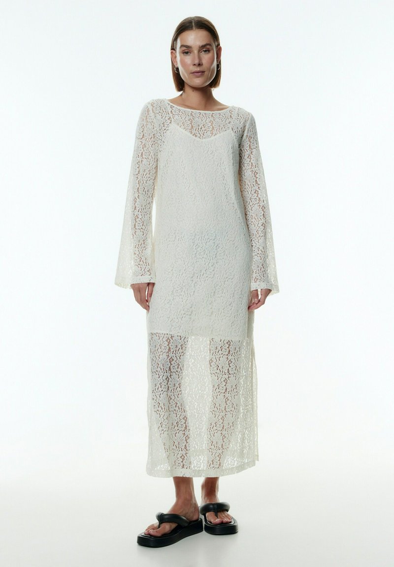 Weißes Spitzenkleid mit langen Ärmeln, transparenter Textur und hohem Ausschnitt. Verfügt über einen mittellangen Saum und ein geschichtetes Design. Schwarze Sandalen runden den Look ab.