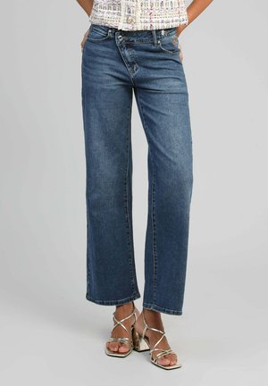 Femme portant un jean bleu taille moyenne à jambes larges, associé à des sandales argentées à talons hauts et à lanières, sur un fond uni.