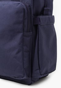 Tamsiai mėlyna kuprinė iš tekstūruotos medžiagos, su keliomis užtrauktukais užsidarančiomis kišenėmis ir šoniniais skyreliais papildomai saugojimui.