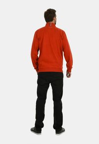 Roter Sweatshirt mit hohem Kragen, der an der Halslinie mit einem horizontalen Akzentstreifen verziert ist. Kombiniert mit schwarzen Hosen und grauen Turnschuhen.