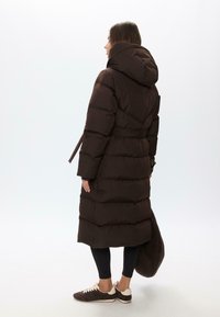 Smeđi jakna s kapuljačom, sa remenom u struku i kviltanom teksturom. Uparen s crnim tajicama i tamnim tenisicama, drži smeđu torbu.