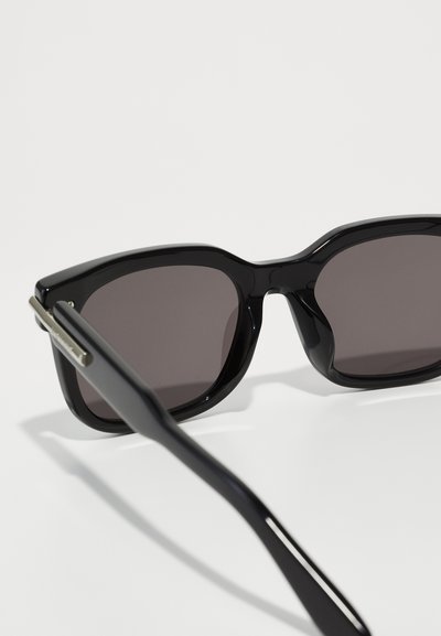 Ochelari de soare negri cu un finisaj lucios, rama unghiulară și lentile întunecate. Accentele metalice de pe brate îmbunătățesc aspectul elegant al designului.