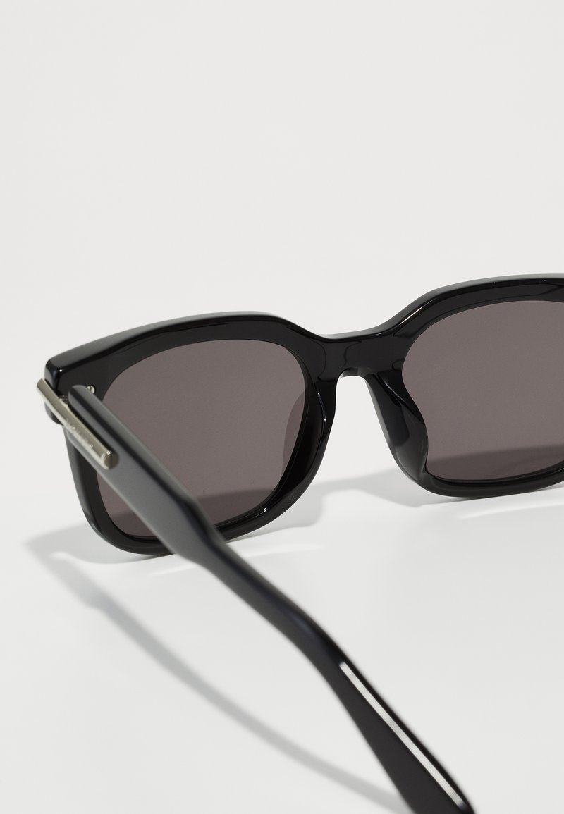 Lunettes de soleil noires avec une finition brillante, des montures angulaires et des verres foncés. Des accents métalliques sur les branches soulignent le look élégant du design.
