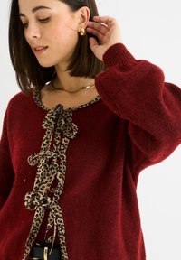 Žena v červeném svetru s leopardím potiskem vpředu, se zlatými kruhovými náušnicemi, tenkým zlatým náhrdelníkem a černým pásem se zlatou sponou.