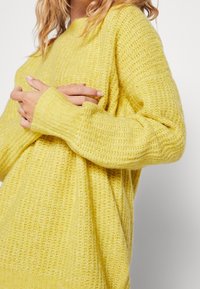 Žena v volném, strukturovaném žlutém pleteném svetru s dlouhými ruk�ávy, se založenýma rukama na úrovni hrudi na jednobarevném pozadí.