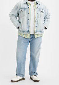 Giubbotto di jeans chiaro con tasche sul petto, indossato su una camicia a righe verde e gialla con colletto, abbinato a jeans a gamba larga e scarpe bianche.