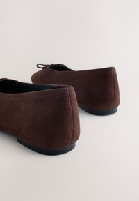 Ballerines en daim marron avec petits nœuds, vues de dos sur un fond clair uni.