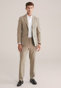 Homme portant un costume beige avec une chemise blanche et des mocassins marron se tenant devant un fond uni clair.