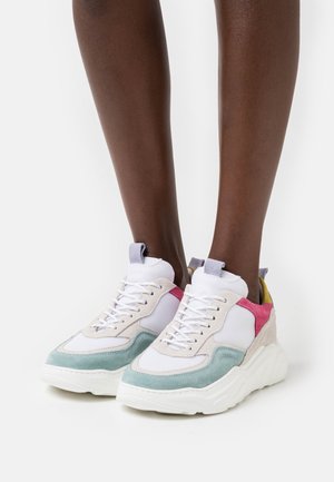 Voeten die chunky witte sneakers dragen met pastelblauwe, roze en gele suède panelen, witte veters en dikke witte zolen tegen een witte achtergrond.