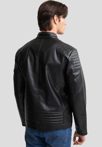 Zwarte leren jas met een hoge kraag, gewatteerde schouderaccenten en geribbeld detail op de onderrug. Draagt met blauwe jeans.