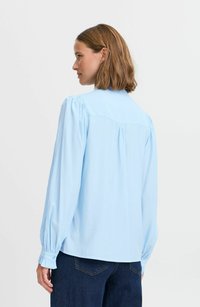 Hellblaues Langarmbluse mit gepufften Schultern, geknöpften Manschetten und geschwunghem Saum. Glatte Textur und geraffte Rückendetails.