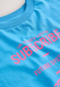Tricou albastru din bumbac cu text imprimat roz: „SUB5CRIBE,” „LDN. 51.5228° N 0.0777° V” și „FUTURE SYSTEM.” Detaliu cu guler striat.