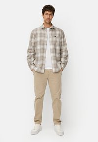 Karierte Hemdjacke in Beige und Weiß über einem weißen T-Shirt, kombiniert mit beigen Hosen und weißen Turnschuhen. Legerer Schnitt und sichtbare Textur.