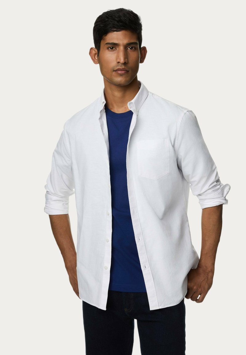 Weißes Hemd mit aufgerollten Ärmeln über einem blauen T-Shirt. Mit einer Brusttasche und einer lässigen Passform. Getragen mit dunklen Jeans.