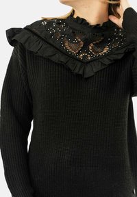 Maglione nero con un corpo a maglia testurizzata e un colletto decorativo nero con volants, caratterizzato da motivi floreali ricamati e dettagli in perline.