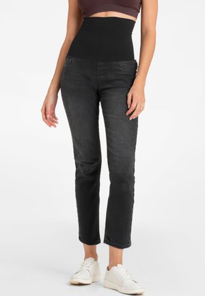 En person iført sorte jeans med høj talje, en crop top i brun farve og hvide sneakers, stående mod en hvid baggrund.