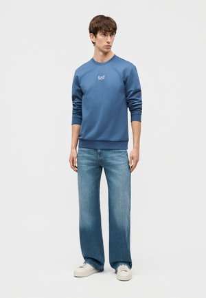 Młody mężczyzna w niebieskiej bluzie EA7, jasnoniebieskich szerokich dżinsach i białych trampkach, stojący na tle jednolitego jasnego tła.