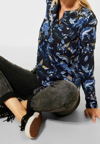 Camicia floreale di un blu navy scuro con motivi blu e gialli, maniche lunghe, colletto, abbinata a jeans grigi e sneakers nere.