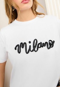T-shirt en coton blanc à manches courtes, avec le mot "Milano" brodé en noir dans une police ondulée et texturée sur la poitrine.