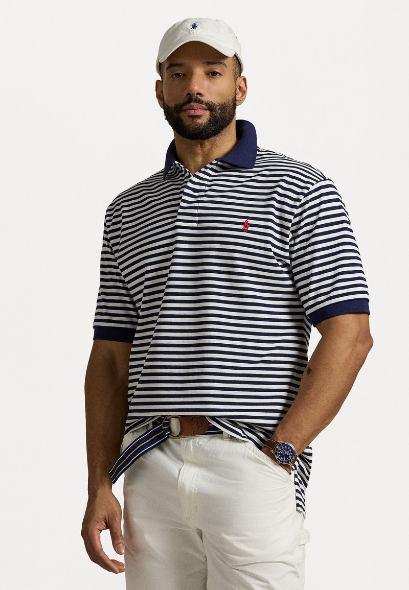 Marineblå og hvitstripete polo-skjorte med marineblå krage og rødt logo. Brukt med lyse bukser og et brunt belte. Hvit caps og klokke synlig.