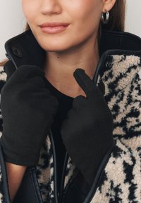 Fekete polár kesztyű, amelyet kézen viselnek, közelről látható egy személy arca és egy mintás külső ruhadarab gallérrészlete. Sima textúra, szoros illeszked�és.