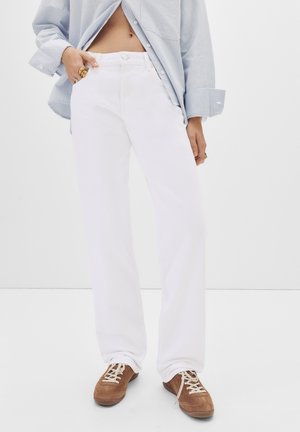 Personne portant un pantalon blanc ample, une chemise rayée bleu clair, des bagues aux doigts et des chaussures en daim marron à lacets sur fond blanc.