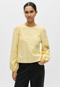 Blouse jaune brodée avec manches longues bouffantes, encolure ronde et motif floral, associée à un pantalon noir. Texture lisse, coupe ajustée.