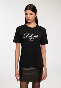 Czarna bawełniana koszulka z krótkim rękawem, z białym, pisanym ręcznie napisem "Delikatny" i graficzną wstążką poniżej. Luźny krój.
