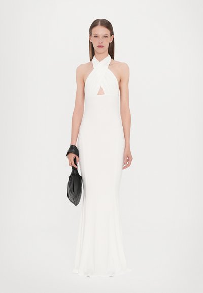 Vestido blanco de cuello halter con silueta ajustada, hecho de tela suave, que presenta un escote en forma de llave en la parte delantera y un dobladillo acampanado. La modelo sostiene un bolso negro.